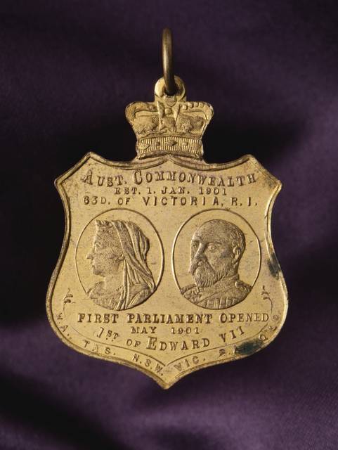 A gold coloured, shield-shaped, Australian Commonwealth Federation souvenir medallion with a crown and suspender ring at the top. In the centre of one side is a portrait of the Duke & Duchess of Cornwall and York. Text above reads 'OPENING OF THE FIRST / FEDERAL PARLIAMENT'. Text below reads 'T.R.H. / THE DUKE & DUCHESS OF CORNWALL / AND YORK / MELBOURNE, MAY, 1901'. On the opposite side is a portrait of Queen Victoria and Edward VII. Text above reads 'AUST. COMMONWEALTH / EST. 1. JAN. 1901 / 63D. OF VICTOR