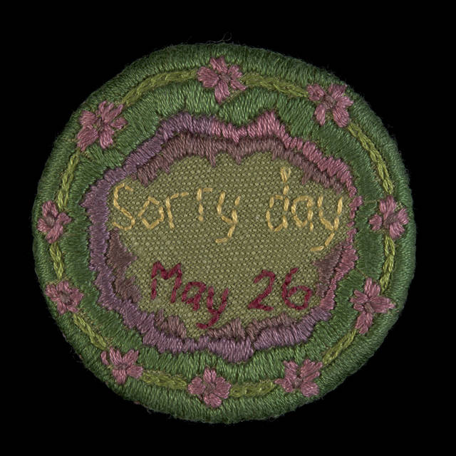 A round badge with the words 'Sorry day / May 26' embroidered in yellow and maroon at the centre, encircled by a decorative purple and pink border, with pink and purple native hibiscus flowers around the outer circumference. The background is green, darker around the outer edge and paler in the centre.The back of the badge is covered with a green cloth and has an attached metal pin.