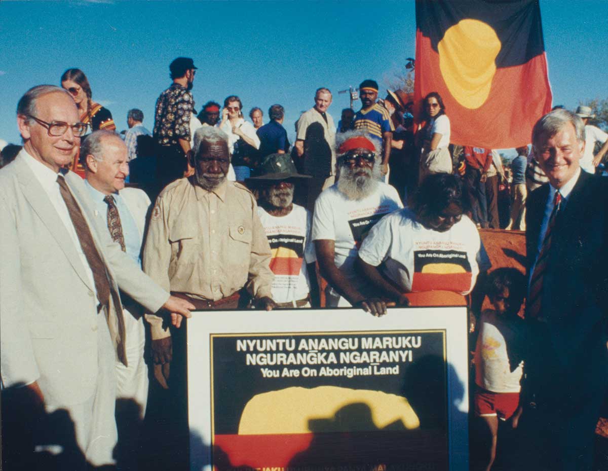 <p>Traditional owners Mr Peter Bulla, Mr Peter Kanari, Mr Nipper Winmarti and his wife, Barbara Tjirkadu with Sir Ninian Stephen, Mr Holding and Mr Cohen (extreme right) and the special poster marking the handback of the title to Ulu<u>r</u>u-Kata Tju<u>t</u>a National Park.</p>
