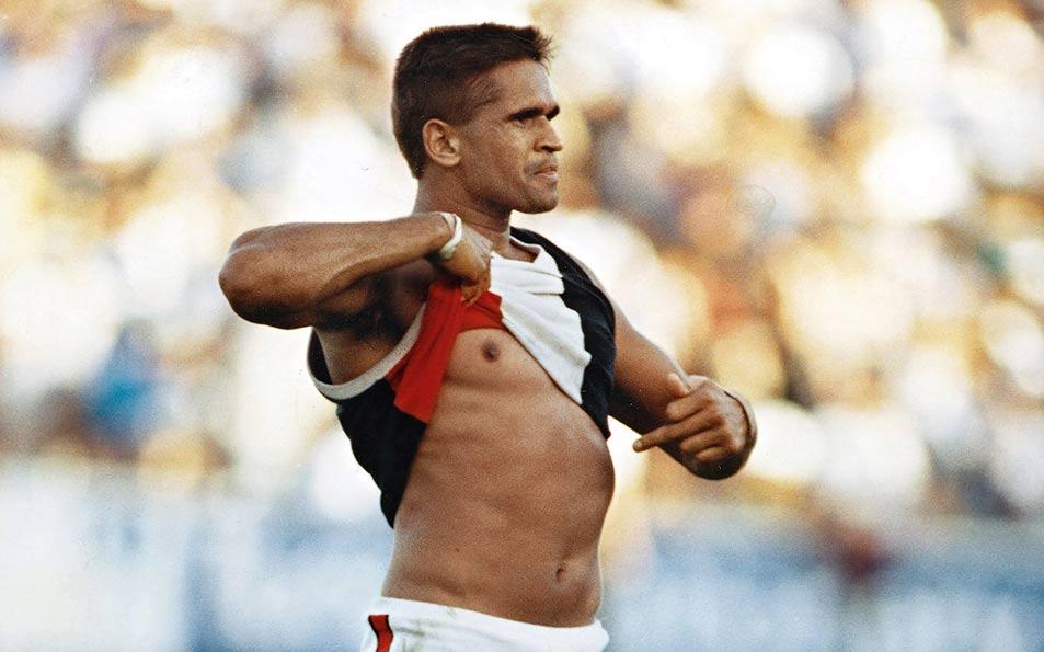 Full length colour photo of an First Nations AFL player standing on the field, lifting up his jumper with one hand and pointing at his belly with the other. He has a determined and defiant expression on his face.