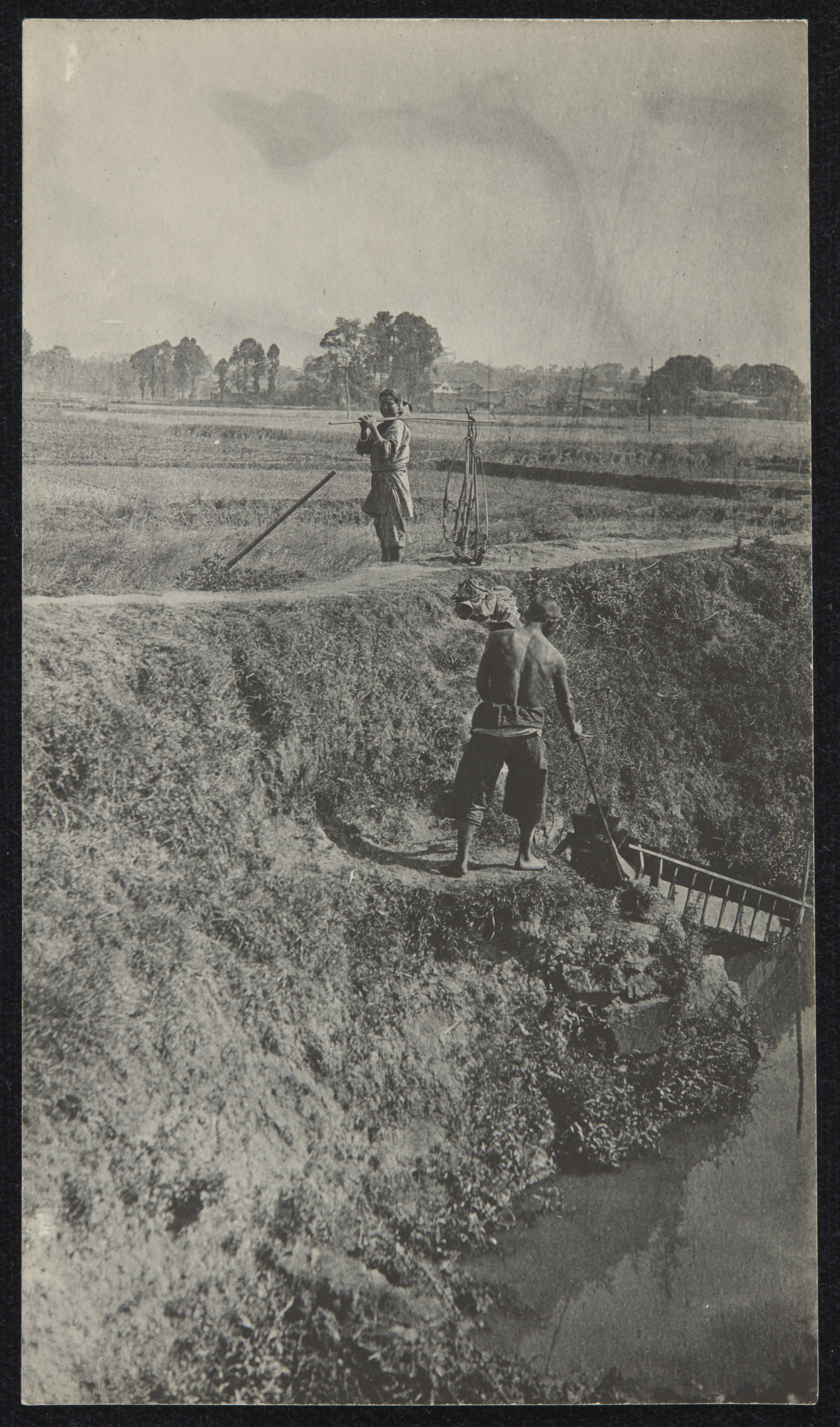 A black and white photograph of two labourers working in a field with water and irrigation devices in the lower right corner of the photograph, 1920.