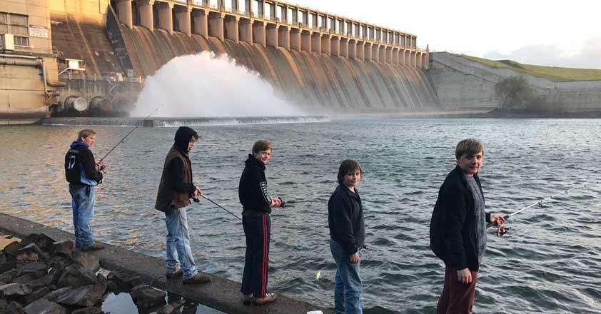 Five teenagers are holding fishing rods and standing on a cement ledge of a dam.