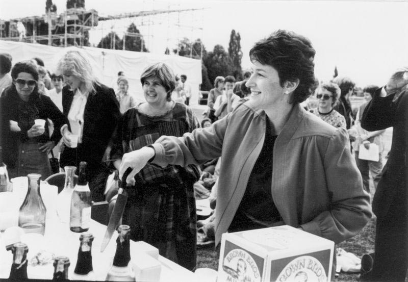 Black and white photograph of a woman smiling at outside event. Several women are in the background. Senator Ryan is holding a knife for cutting a cake. Champagne bottles are visible.