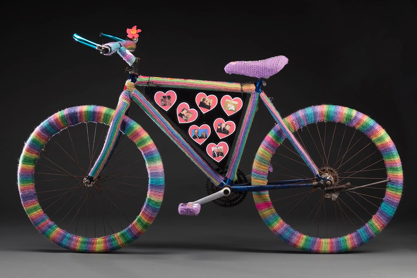 A bicycle covered with woollen crochet that consist of multi-coloured striped wheels, a purple seat and two purple peddles.