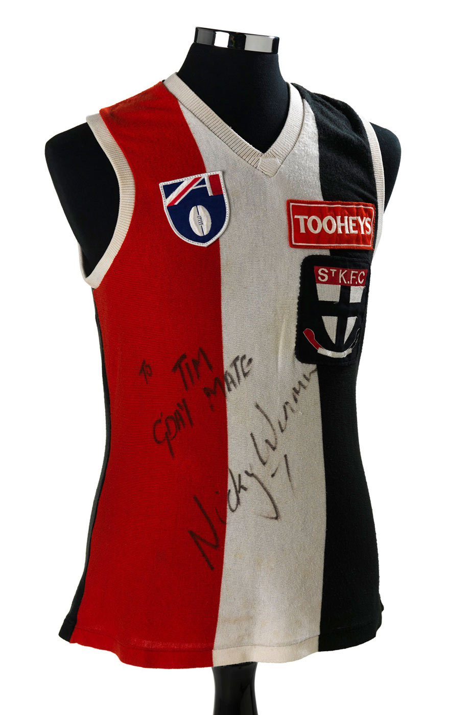 Sleeveless top with vertical red, white and black stripes. There is a St Kilda logo on the breast with the word ‘Tooheys’ above that. The words ‘Tim G’day Mate’ is written in texta and signed by Nicky Winmar.