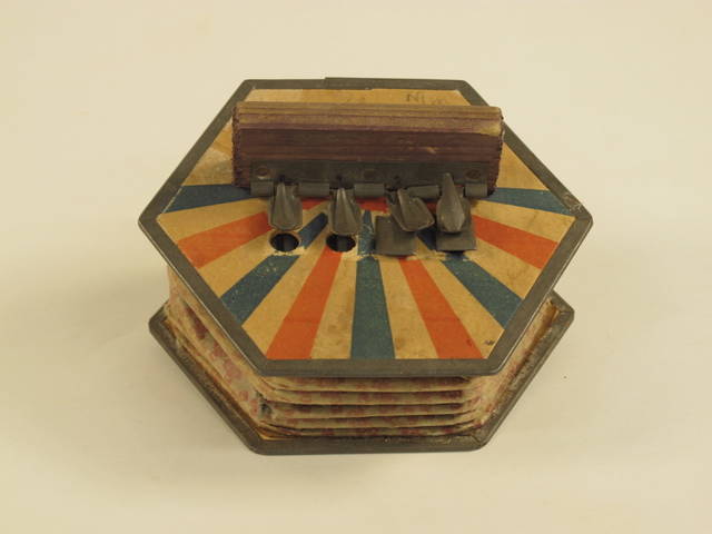 A paper and metal hexagonal shaped concertina with wooden handles and corroded white metal buttons that is decorated with a red and blue rising sun and polka dot motif.