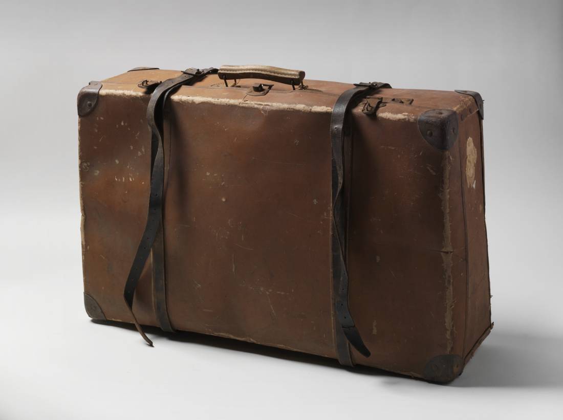 A brown suitcase featuring a wooden handle attached to the top by metal clips, leater corner protectors and metal clasps a a pair of brown and black leather and metal suitcase straps.