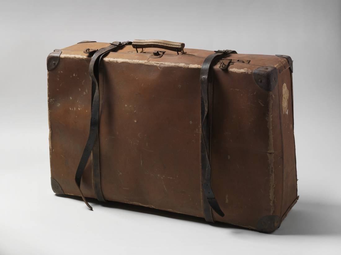 A brown suitcase featuring a wooden handle attached to the top by metal clips, leater corner protectors and metal clasps a a pair of brown and black leather and metal suitcase straps.