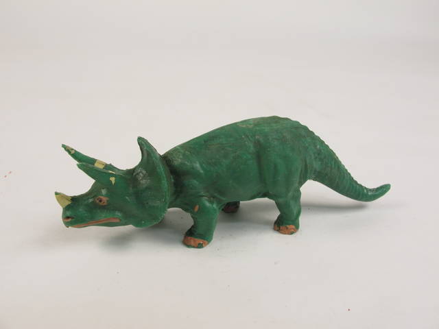 A miniature green plastic toy triceratops with painted facial features, including black and pink eyes, pink nostrils and a mouth.