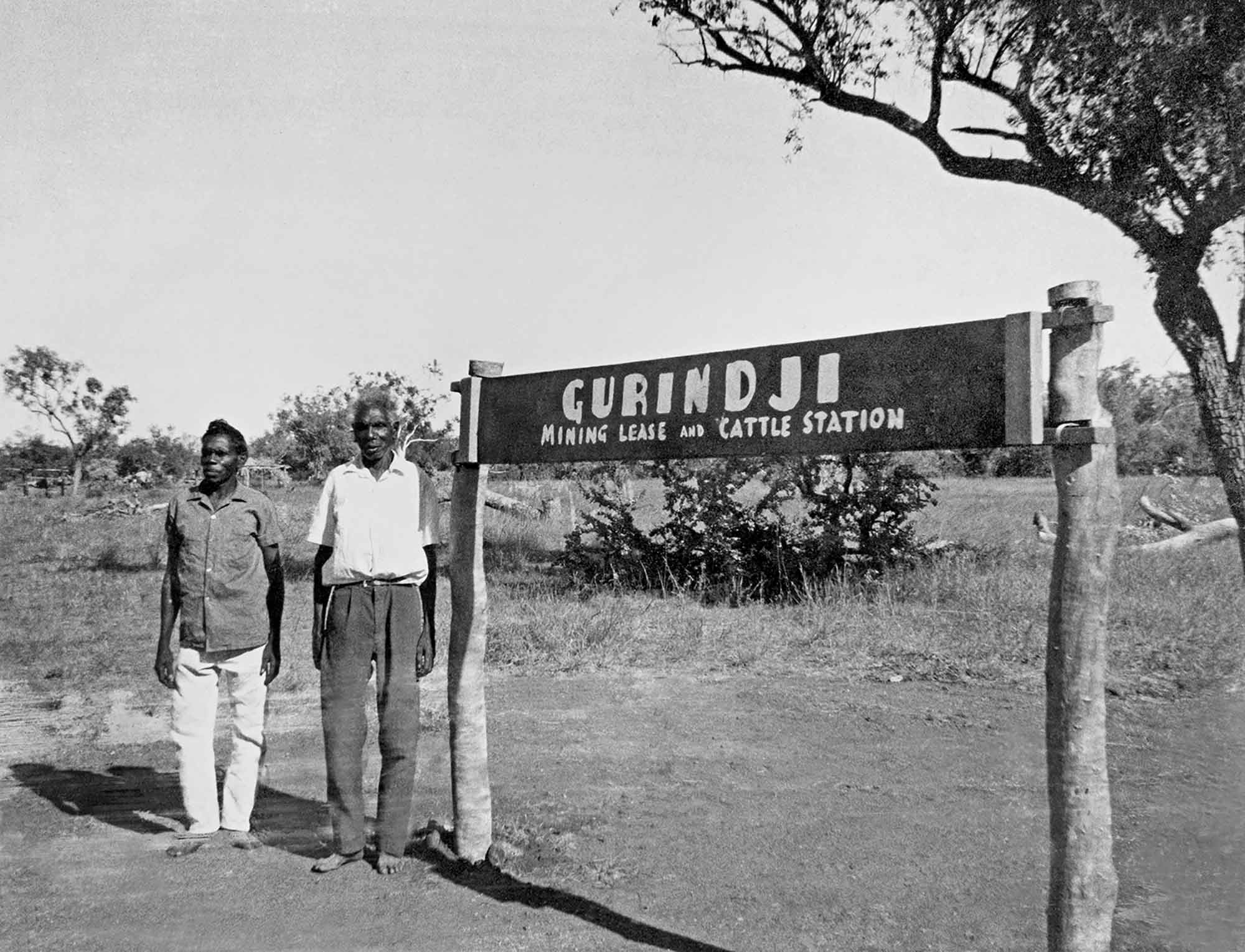 Black and white photo of two men beside a sign reading ‘Gurindji mining lease and cattle station’. 