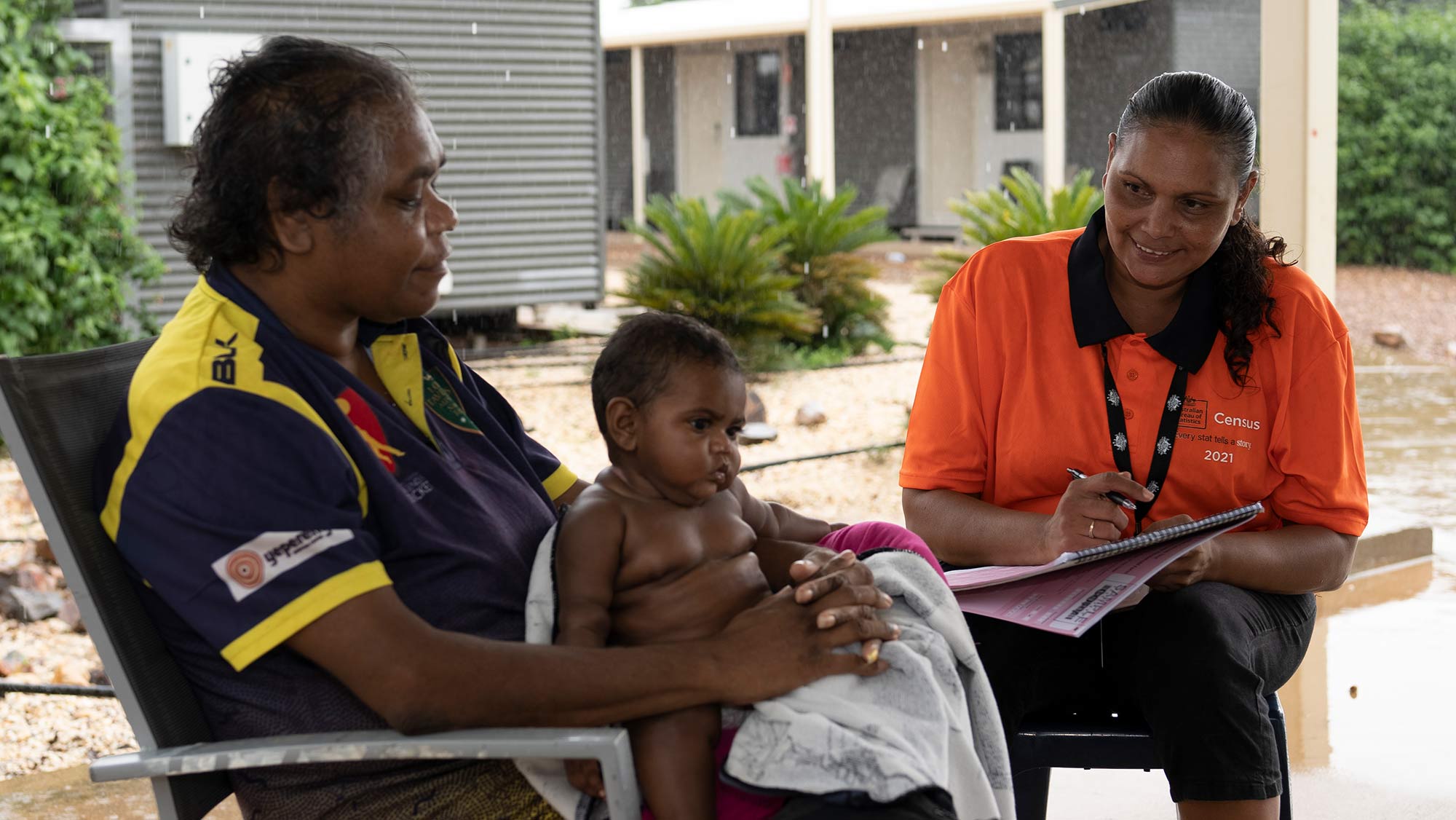 A woman is sitting in a chair with a child in her lap. A female Census staff member is smiling at the child with a pen and log book in her lap.