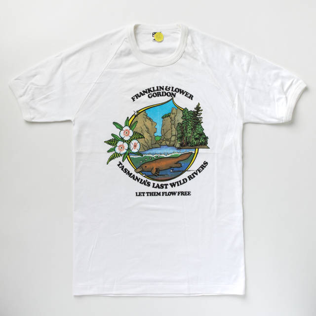 A white t-shirt with a colourful Wilderness Society emblem on the front which features a platypus with flowers, trees and a rocky gorge inside a tear drop shape. Black text above and below the emblem reads "FRANKLIN & LOWER GORDON TASMANIA'S LAST WILD RIVERS...LET THEM FLOW FREE." The manufacturers label reads 'BONDS.16'.
