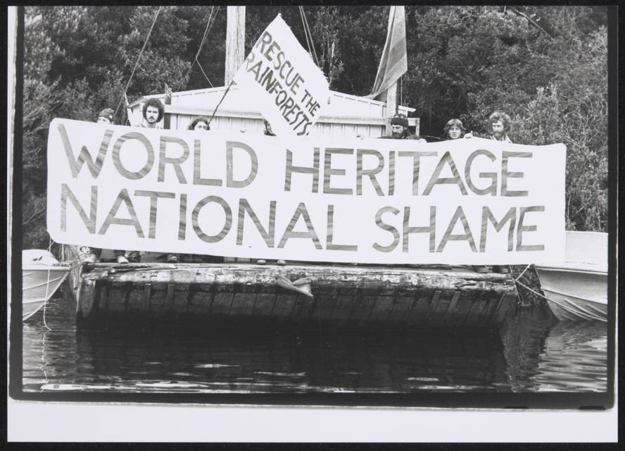 A black and white photograph of seven protesters standing on a jetty holding a large banner that reads "WORLD HERITAGE / NATIONAL SHAME". A smaller flag is being waved by one protester that reads "RESCUE THE RAINFORESTS".