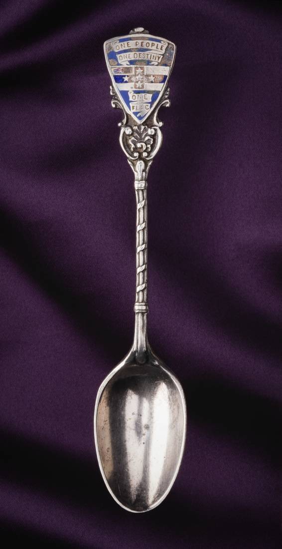 A silver souvenir teaspoon with a chipped blue enamel shield at top of handle. The shield features part of Union Jack and a horizontal and vertical row of three stars forming a cross. Text in ribbons within shield read 'ONE PEOPLE / ONE DESTINY / ONE FLAG'. The back of the shield is hallmarked with a crown, lion, letter 'g' or 'q', and a pennant. 'RD. NO. 348138' is impressed in the back of the handle's shaft.