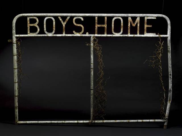 A metal gate panel with welded metal letters at the top which read 'BOYS HOME'. Remnants of wire mesh are attached to the tubular upright components of the gate, which are on the outer edges and in the centre.