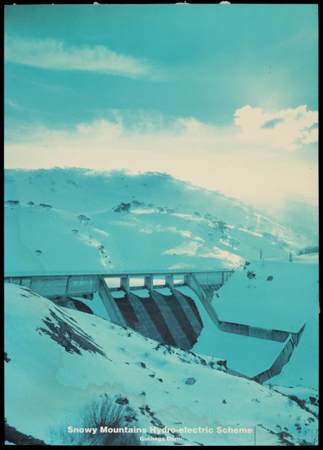 A poster featuring a photograph with the caption 'Snowy Mountains Hydro-electric Scheme / Guthega Dam', mounted on foam board. The pictures shows a dam wall and mountain behind, covered in snow. The picture has faded to a shade of blue, with brown water stains. On the back of the poster there is a small label for George Mico of Hollywood Studio, Cooma. The back also has gouges, and a small metal fitting attached.