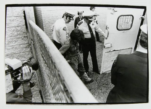 A black and white photograph of a woman in overalls looking at the ground so her face can not be seen. She is being escorted to the back of a police van by two police officers. A third policeman is stranding by the door and a police woman and another policeman can be seen standing in the background. This picture is taken over a fence which can be seen in the bottom left hand side of the picture. Another camera is filming through the fence.