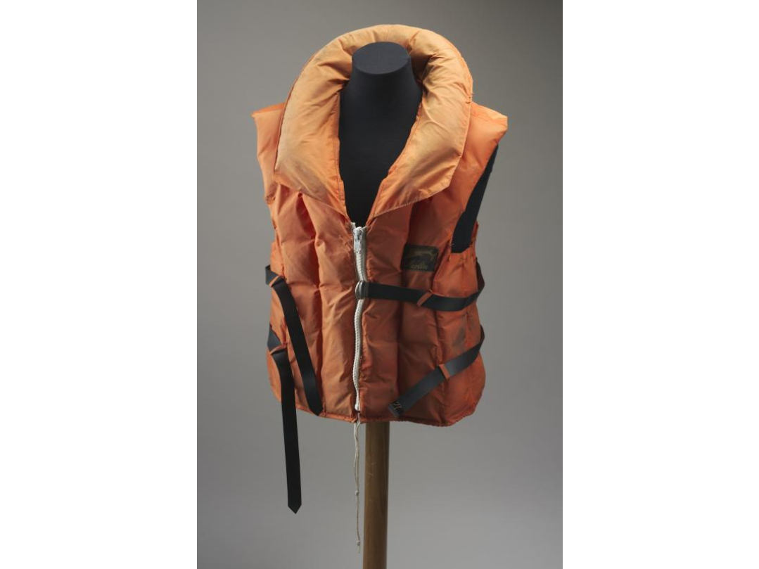 An orange fabric life jacket, padded with plastic air filled pillows. The jacket has a white zip at the front, with two black plastic belts around the chest and waist and a drawstring around the bottom edge to hold it on firmly. A label sewn into the jacket gives a warning and instructions. The manufacturer's label on the front reads 'Marlin'.