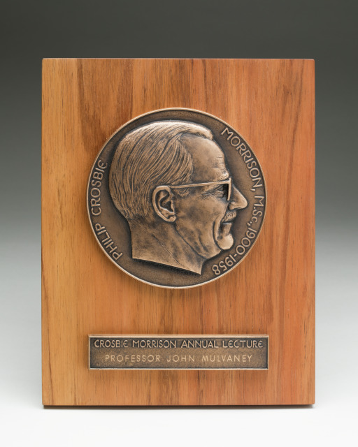 Metal plaque mounted on a rectangular piece of wood. The metal plaque is round and features a man's head in relief and the text "PHILIP CROSBIE MORRISON, M.SC., 1900-1958" around the edge. There is a separate metal rectangle below that reads "CROSBIE MORRISON ANNUAL LECTURE / PROFESSOR MULVANEY".
