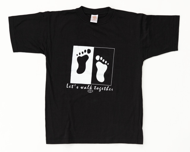Black cotton t-shirt with white printed text which reads "Let's walk together" and a small logo which reads "THIRD EYE / PARTNERS". The image printed on the t-shirt consists of a black footprint in a white rectangle and a white footprint in a black rectangle.