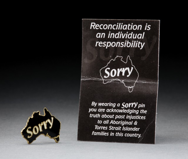 Sorry badge and card produced by Link-Up. The gold coloured metal hat badge is in the shape of an outline map of Australia. It is inlaid with black enamel, and the word 'Sorry' is accentuated with white enamel running across the badge. The accompanying card has white writing printed on a black background. 