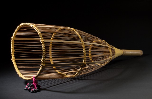 A cylindrical fish scoop made of wood, sticks and yellow string. There are fifteen black seed pods attached with pink wool to one side of the open end of the scoop.