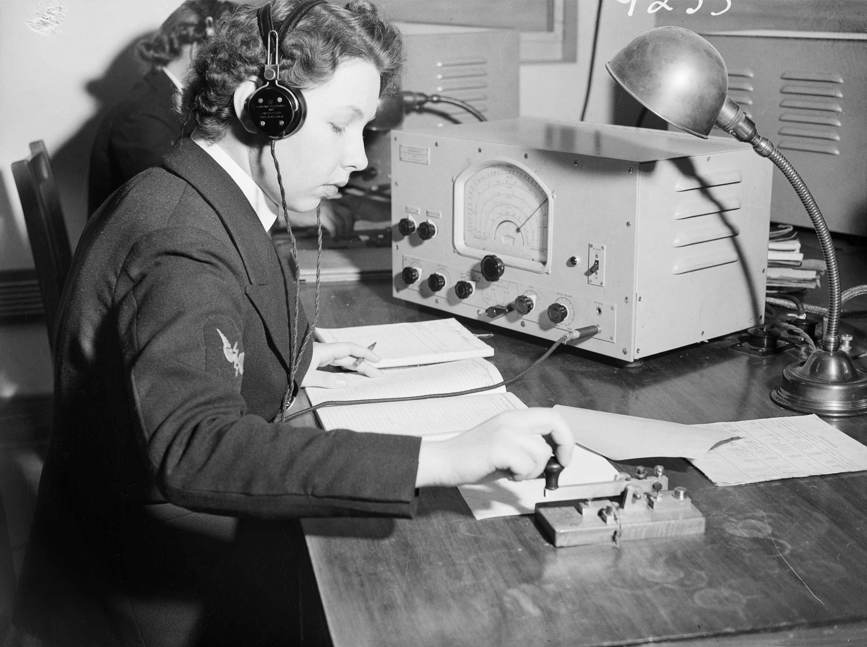 A woman wearing a uniform and headphones works at a desk with radio receiver, morse key, books and a lamp. 