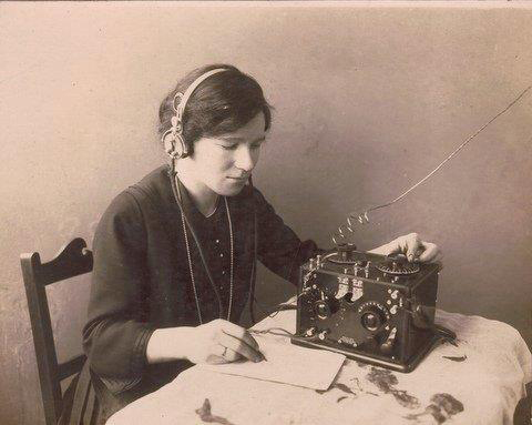 A woman wearing headphones works at a desk with a radio receiver and notepad.