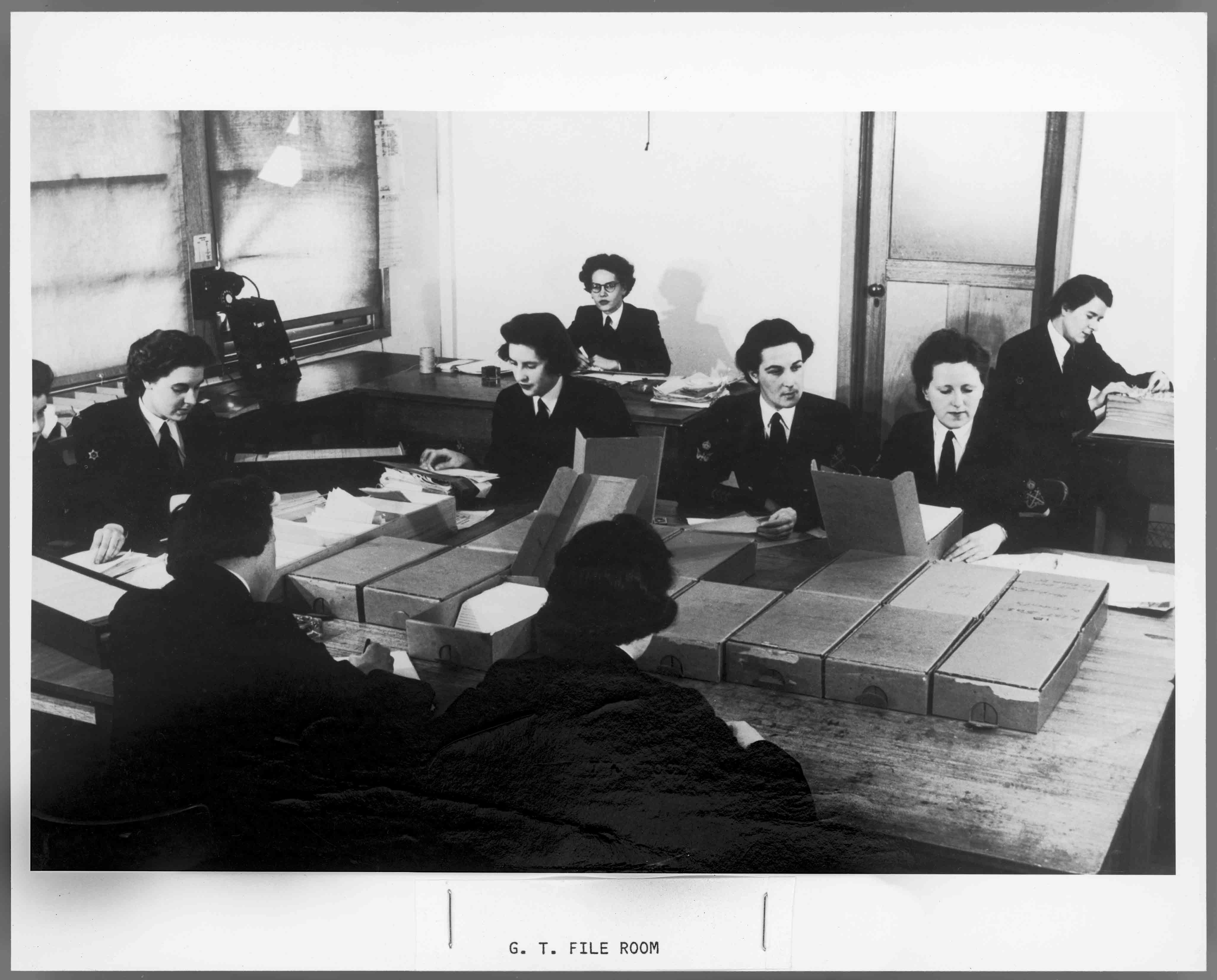 Nine women working at shared desks with paper files and boxes on them. 