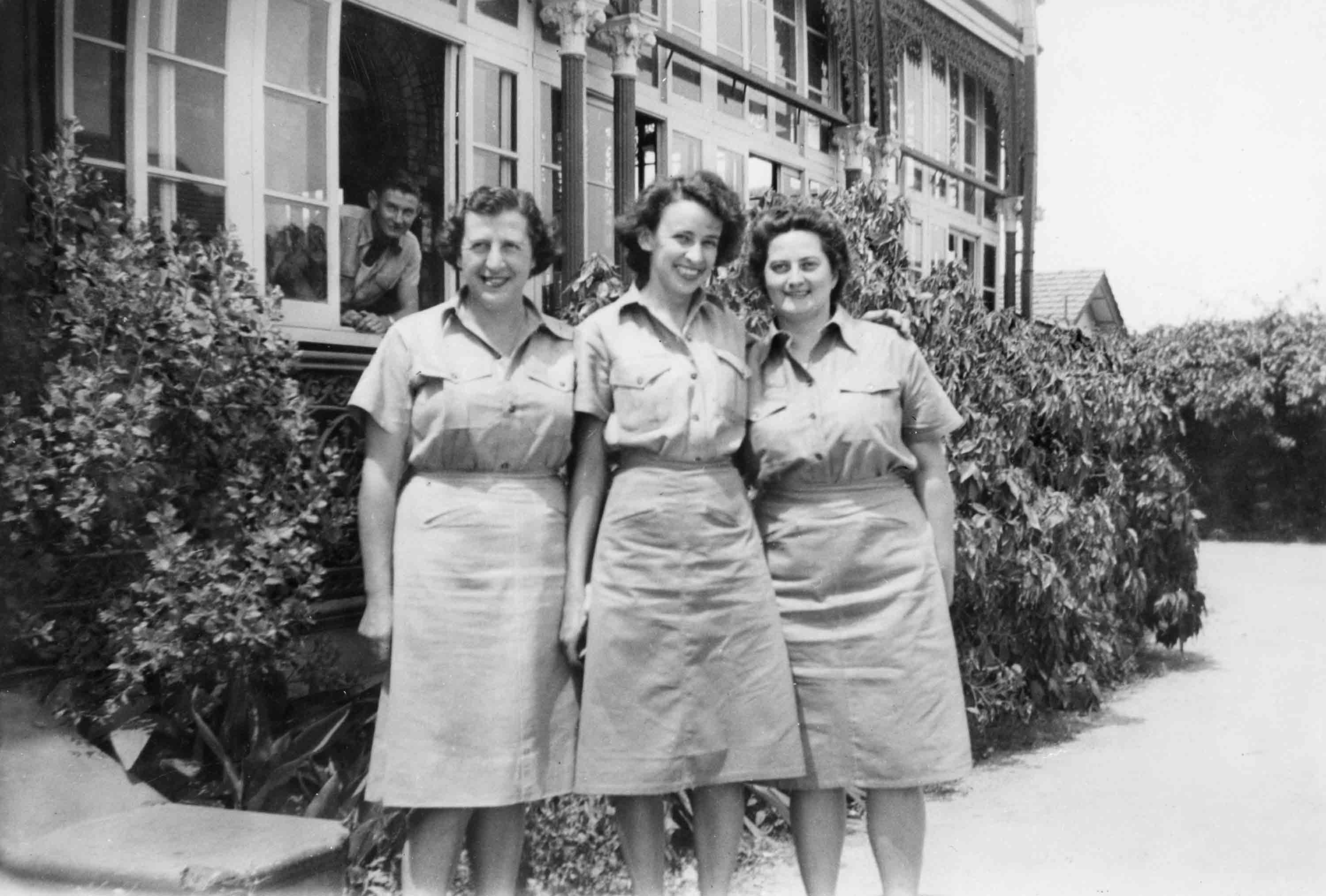 Three women in uniform stand in front of a building smiling. 