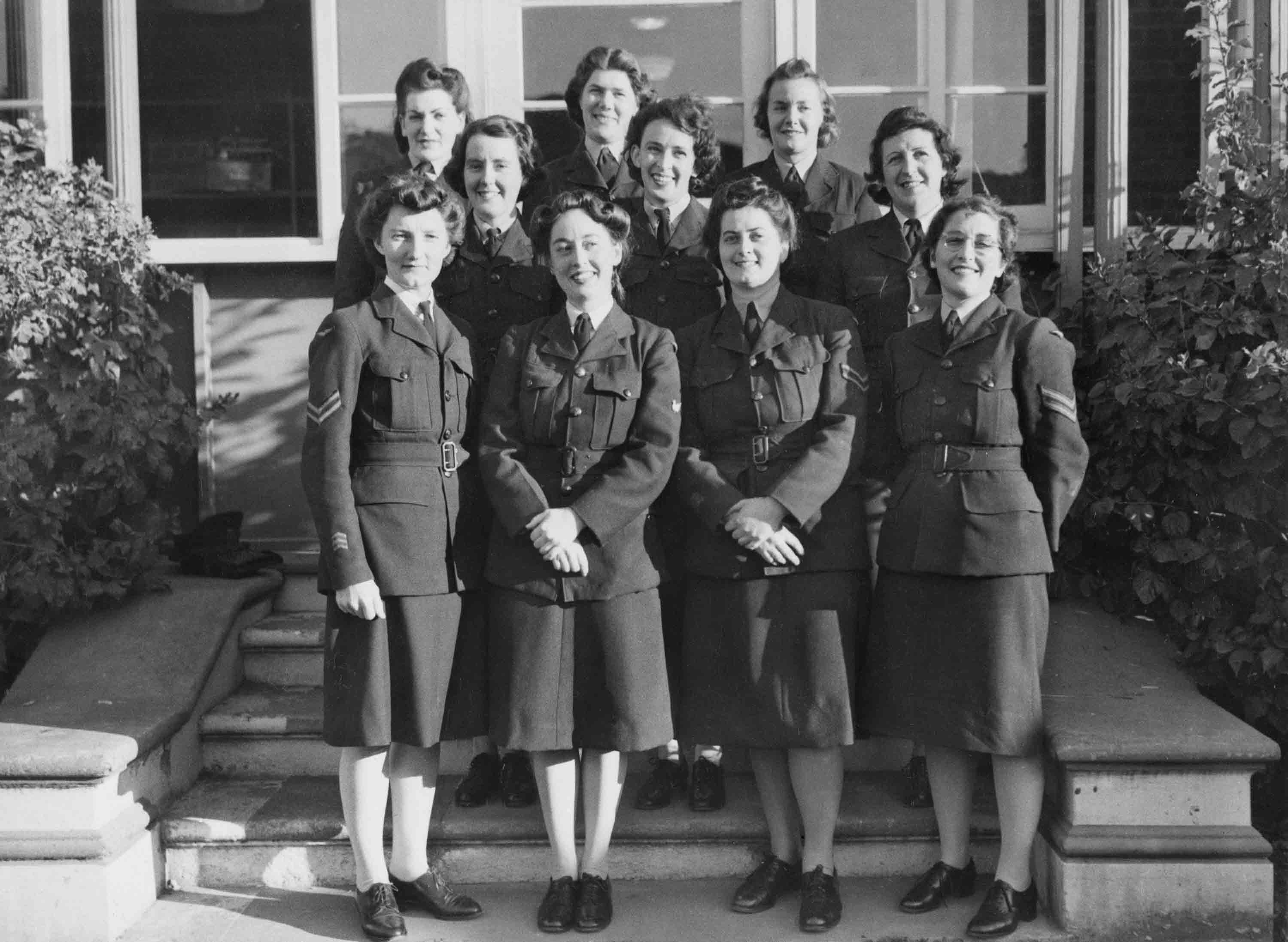 Ten women in uniform standing in three rows on a set of stairs in front of a building.