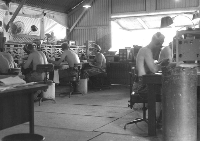 Shirtless men with headphones on work in front on radio receivers in a corrugated iron building.