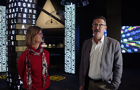 Two people stand in a dark exhibition space with letters and lights behind them.