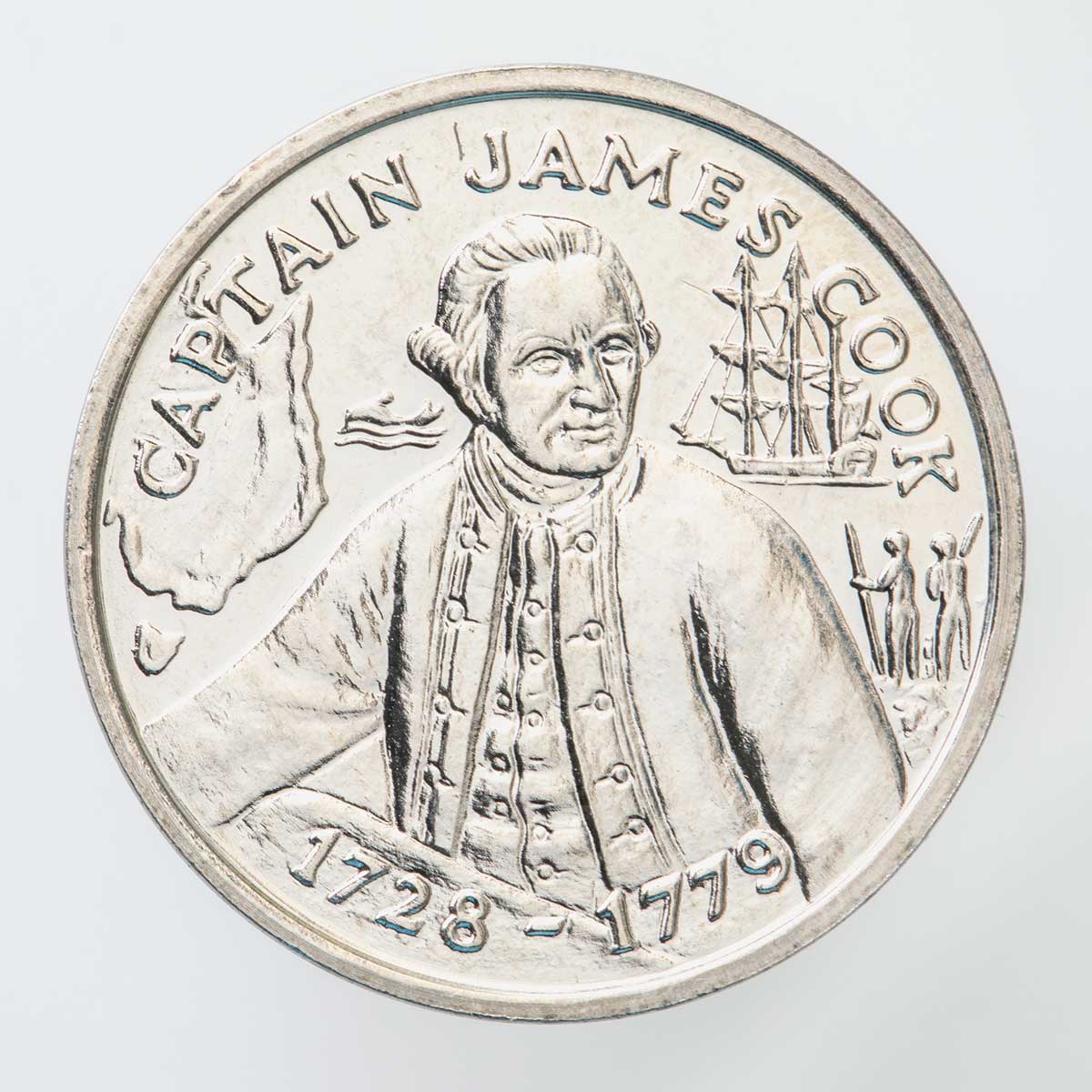 A silver coloured metal commemorative medal depicting Captain James Cook. Smaller images of a ship, Indigenous Australians and the east coast of Australia are depicted on the front, with text stating "Captain James Cook" and dates "1728-1779" inscribed on the medal.