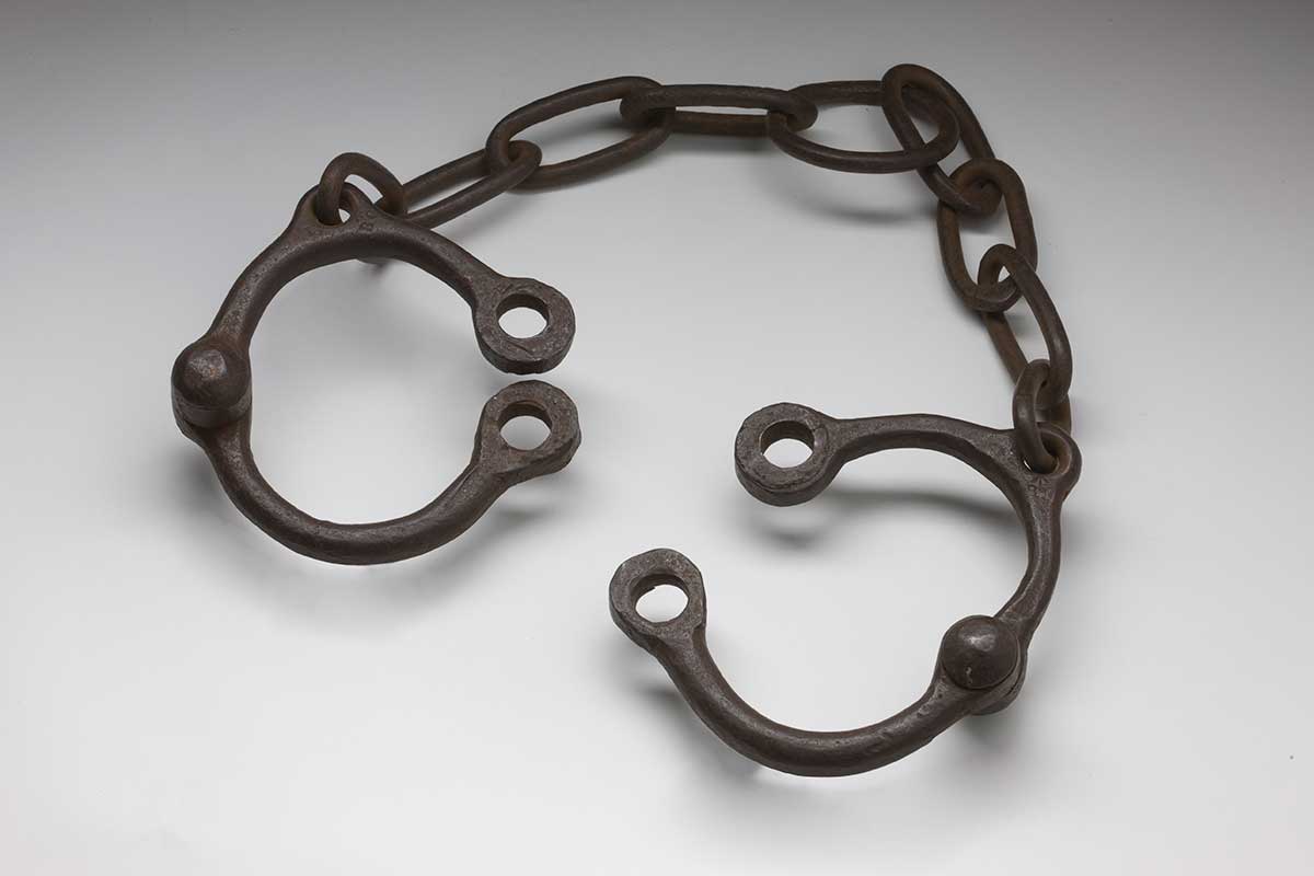 A pair of convict leg irons with leg cuffs that open at a hinged rivet, and are linked to each other by eight oblong links of chain with a circular link at the mid-point.