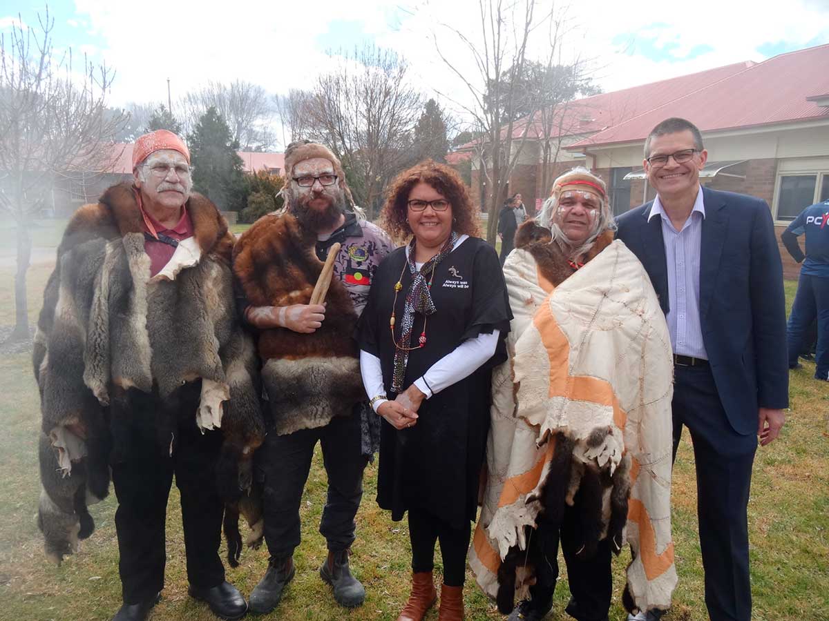 Group portrait of four men and one woman. Three men are wearing possum skins and one man is holding clap sticks.