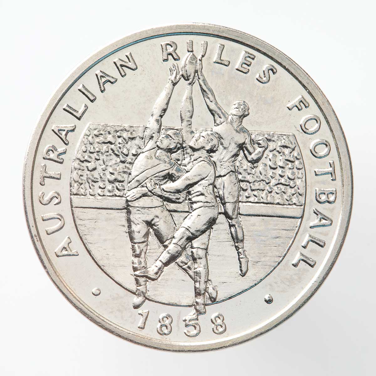 A silver coloured metal commemorative medal depicting Australian Rules Football with three footballers playing in front of a crowd with the text "Australian Rules Football" and the date "1858".
