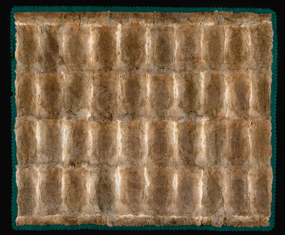 A large, brown coloured, rabbit skin rug that is lined and edged with emerald green felt. The felt edging is scalloped. There are 40 rectangular pelts used in the main section of the rug and several pelts cut into thinner strips that run along the two longer edges of the rug.