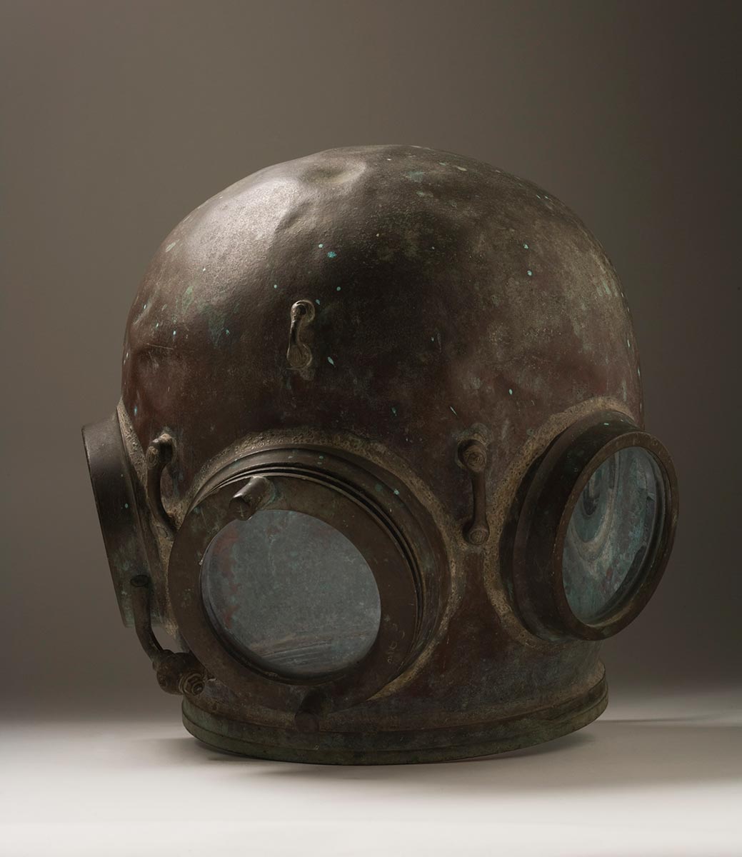 Metal diving helmet featuring a round head shape with round glass windows on three sides.The front window has two straight handles attached to it and there are metal loops welded in various places.