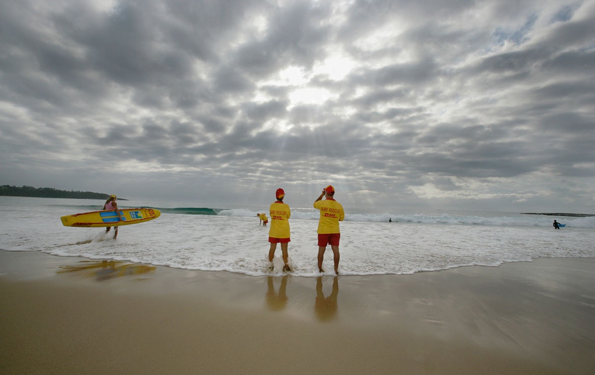 Two life savers stand on the beach looking out towards the surf. Another life saver enters the surf holding a surf board, and another life saver in the distance appears to be searching the water. 