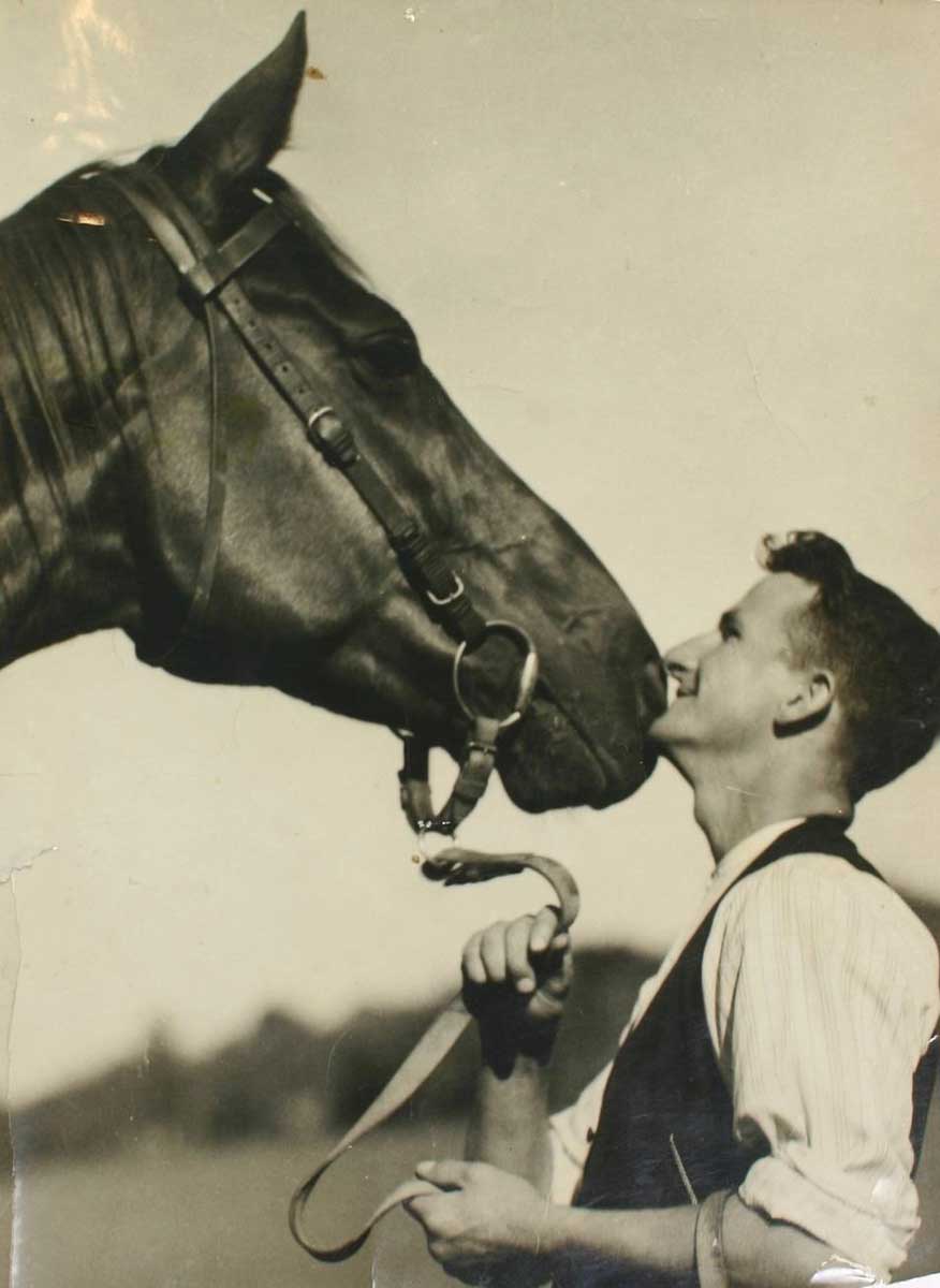 Black and white photo of a man smiling affectionately towards a horse.