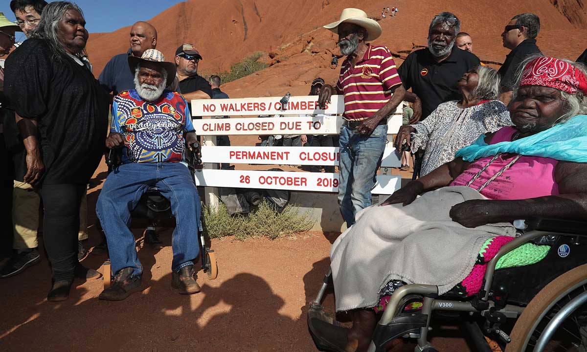 Colour photograph of a group of First Nations peoples gathered at the base of Uluru, around signage banning the climbing of the landmark.