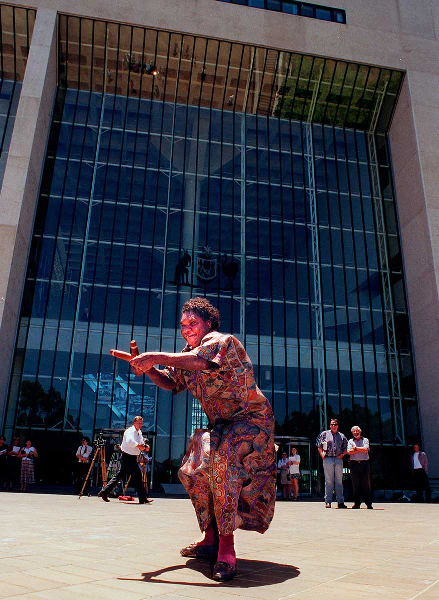 <p>Aurukun Elder Gladys Tybingoompa dances on the steps of the High Court, which had just ruled in favour of the Wik native title claim, 1996</p>