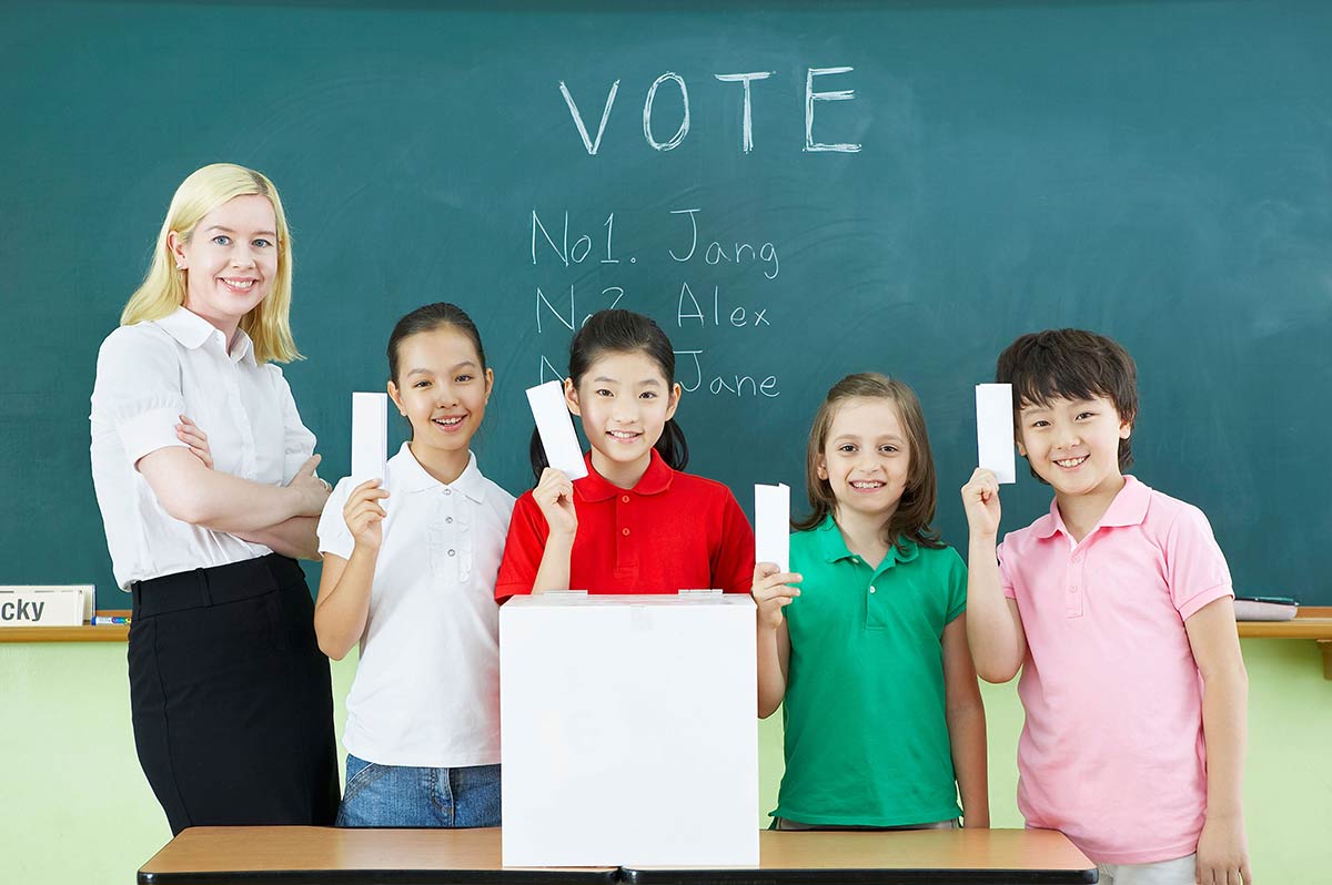 One teacher standing with a group of four young students in front of a chalk board. Written on the board is 'VOTE' with candidates names below. The students are holding ballot papers in the air.