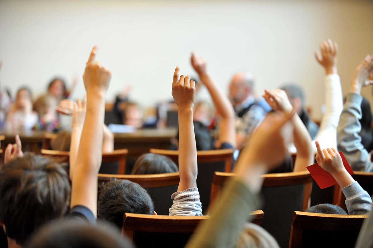 View from behind of children sitting on chairs raising their hands.