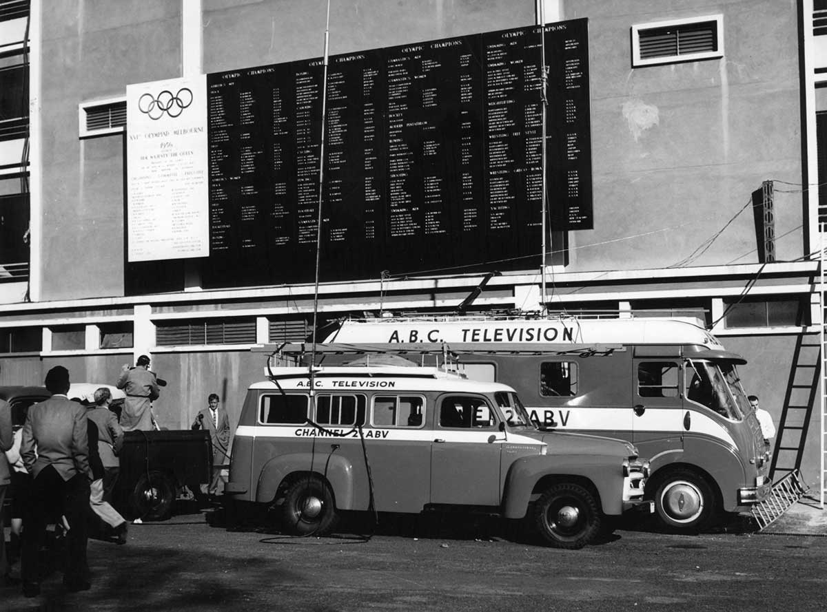 A black and white photograph of two ABC Television broadcast vans stationed below a fixture board featuring the Olympic rings. A press conference appears to be taking place.