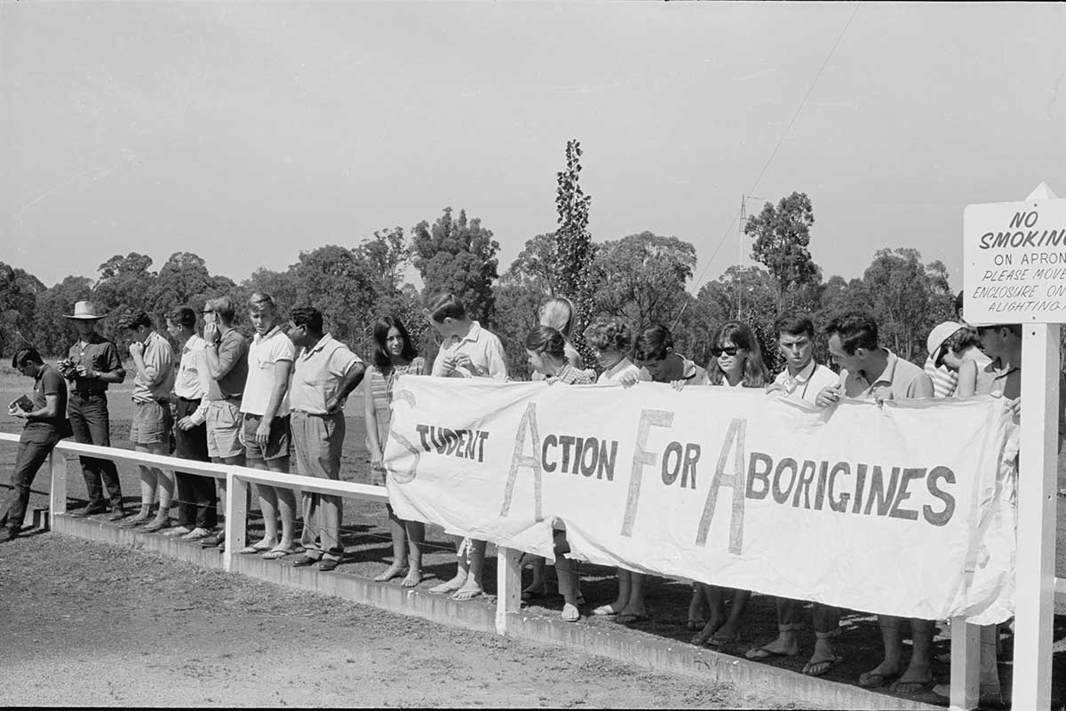 A group of men and women standing on one side of a low fence. Half of them are holding a large banner printed with 'STUDENT ACTION FOR ABORIGINES'.
