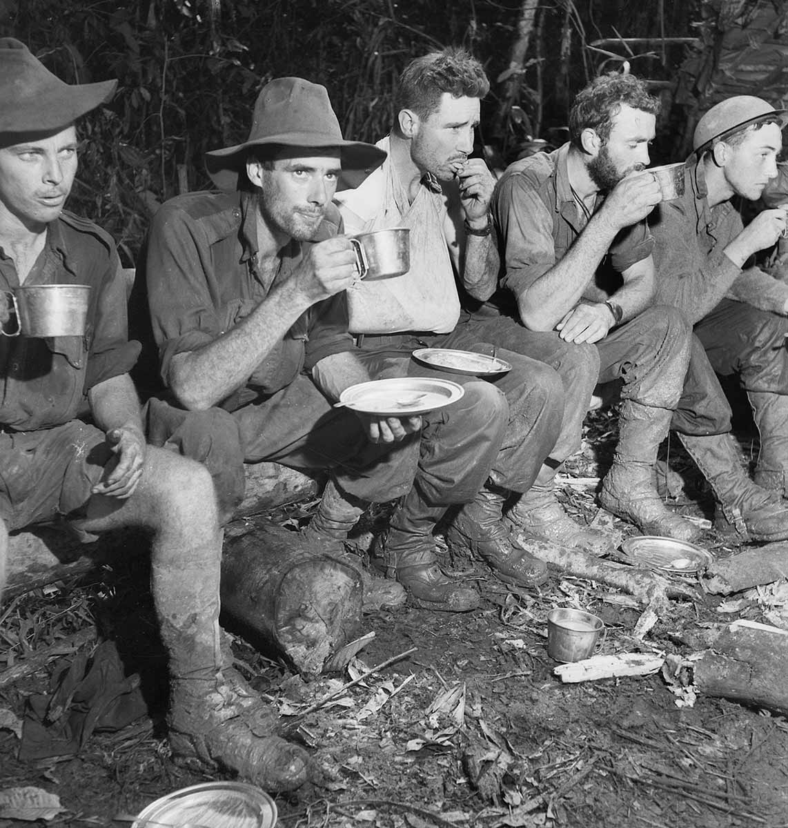Black and white photograph of Australian soldiers resting. They are sitting on a log and eating and drinking. One of the soldiers' arm is in a slingshot.