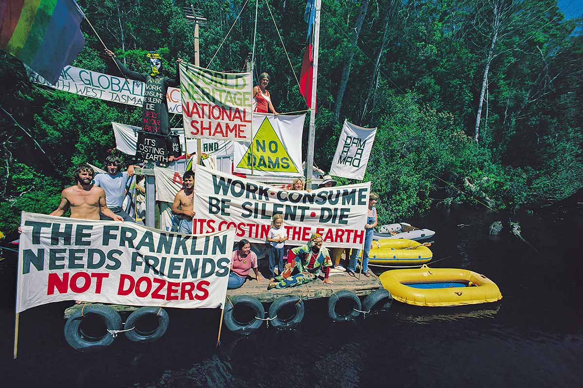 A group of protestors are sitting on rafts on the edge of the Franklin River, holding protest banners.