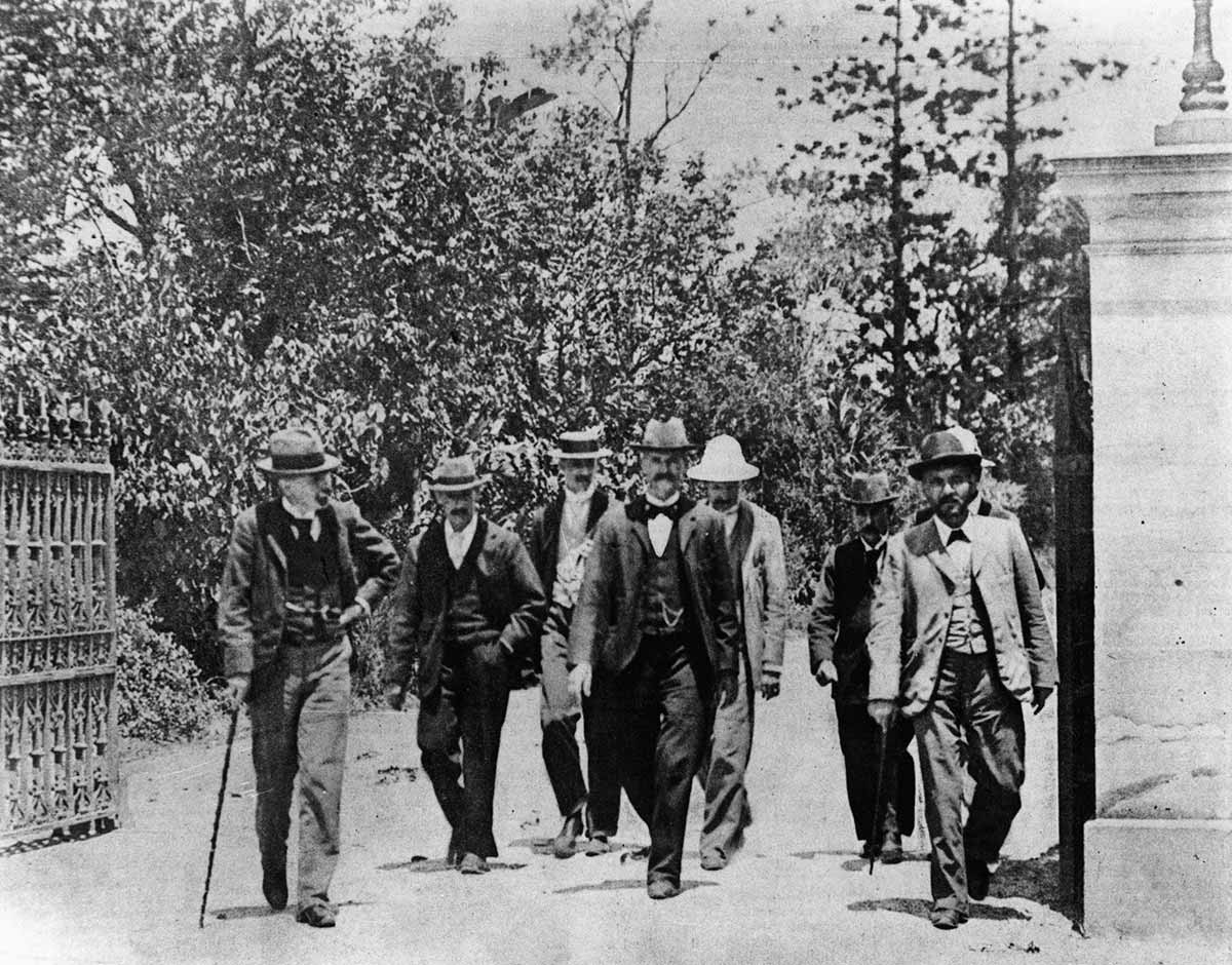 Black and white photograph of 7 or 8 men in double-breasted suits walking leisurely together through ornate driveway gates.